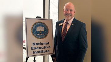 Noel March, wearing a suit and tie, stands beside a sign with a seal that reads, "Federal Bureau of Investigation National Executive Institute"