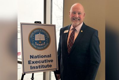 Noel March, wearing a suit and tie, stands beside a sign with a seal that reads, "Federal Bureau of Investigation National Executive Institute"