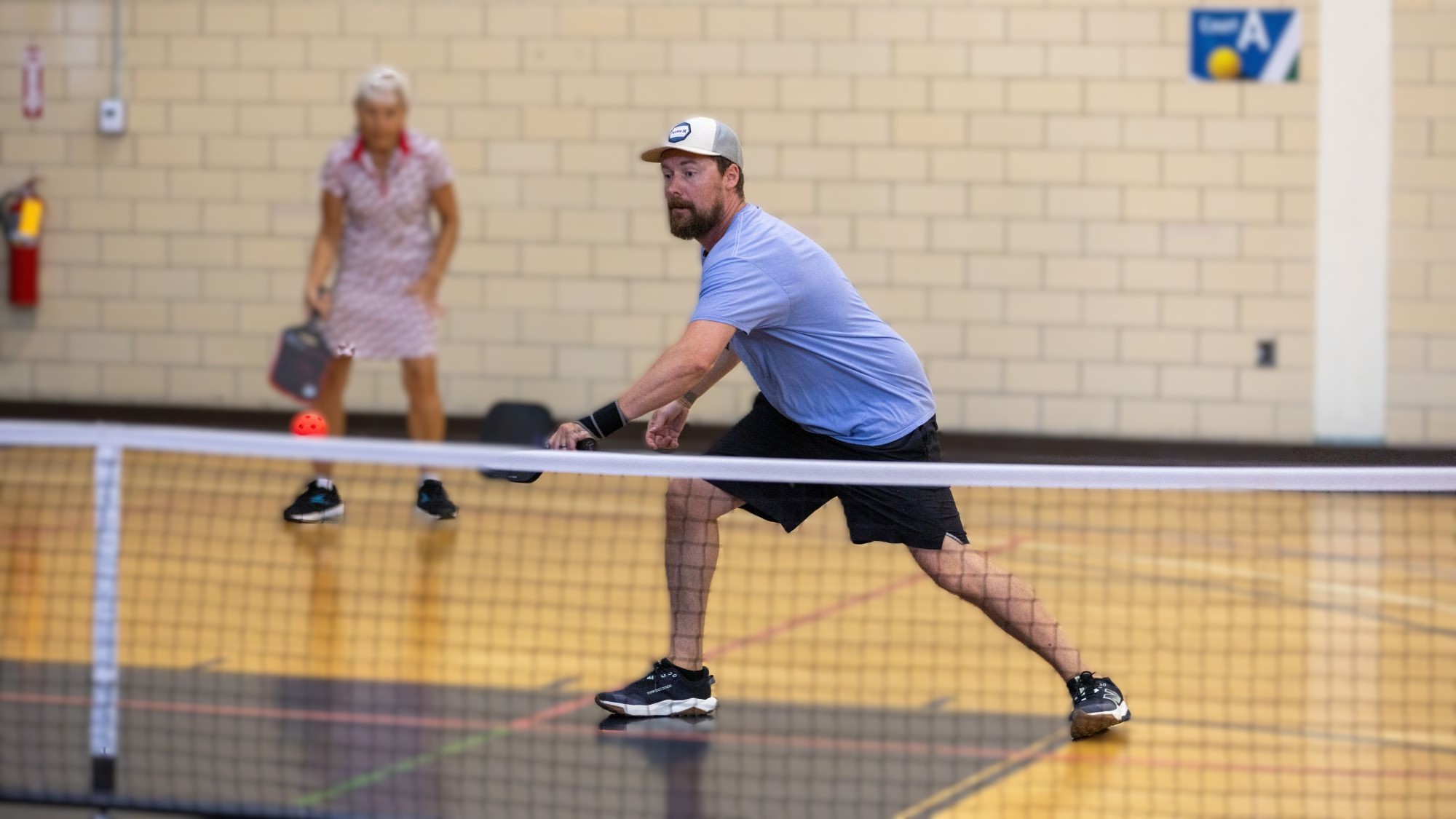 A man wearing a light blue t-shirt lunges to hit a pickle ball with a paddle as a woman in the background stands at the ready.