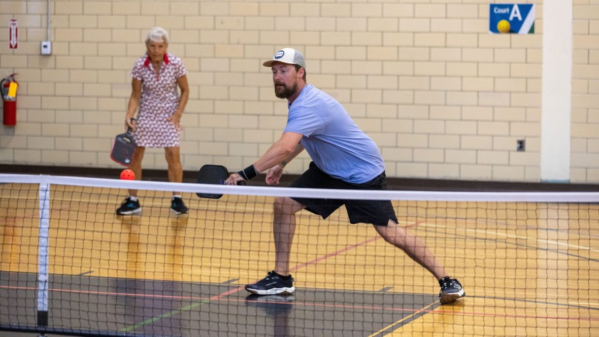 A man wearing a light blue t-shirt lunges to hit a pickle ball with a paddle as a woman in the background stands at the ready.