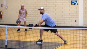 A man wearing a light blue t-shirt lunges to hit a pickle ball with a paddle as a woman in the background stands at the ready.