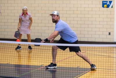 A man wearing a light blue t-shirt lunges to hit a pickle ball with a paddle as a woman in the background stands at the ready.