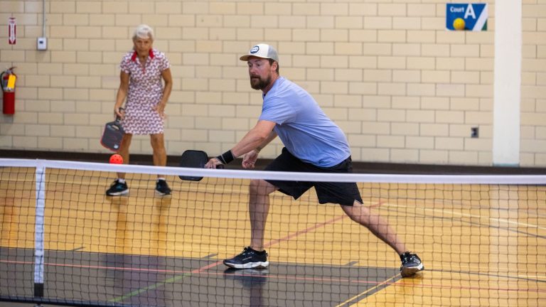 A man wearing a light blue t-shirt lunges to hit a pickle ball with a paddle as a woman in the background stands at the ready.