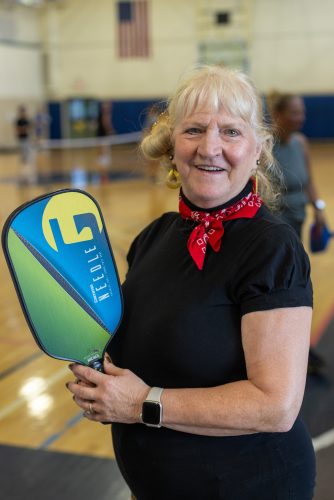 A woman with blond hair, black t-shirt and red bandana tied around her neck smiles and poses with her pickleball paddle while standing in an indoor gymnasium, with pickleball courts and players visible in the background.