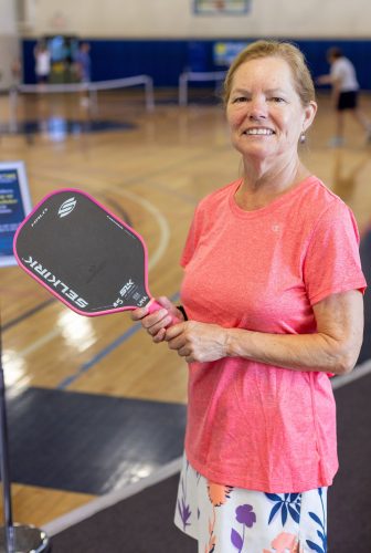 A woman in a bright coral athletic shirt holds a pickleball paddle while standing in an indoor gymnasium, with pickleball courts and players visible in the background.