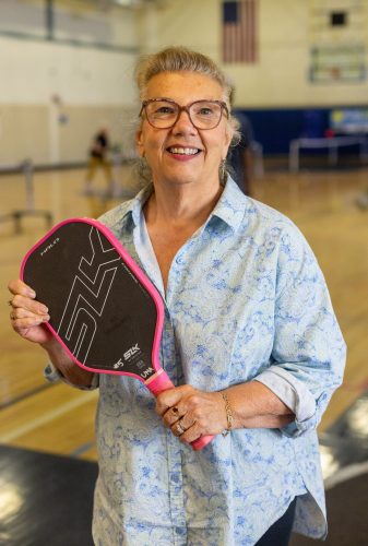 A woman wearing glasses and a button-down shirt poses with her pickleball paddle while standing in an indoor gymnasium, with pickleball courts and players visible in the background.