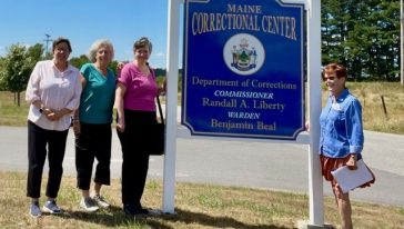 Four CODA Chorus volunteers stand beside the Maine Correctional Center sign in Windham.