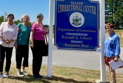 Four CODA Chorus volunteers stand beside the Maine Correctional Center sign in Windham.