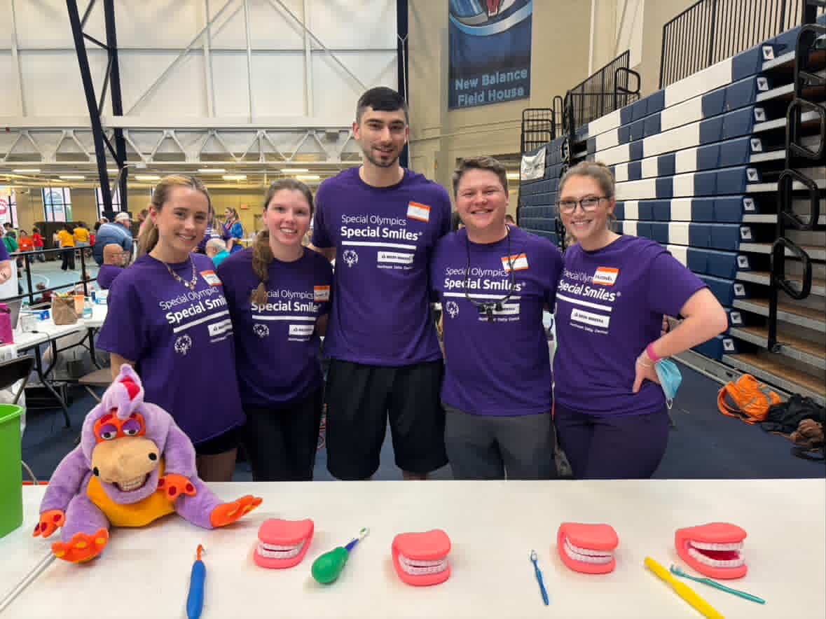 Dental students wearing purple “Special Olympics Special Smiles” shirts pose together at a table with model teeth and dental hygiene tools during a community event.