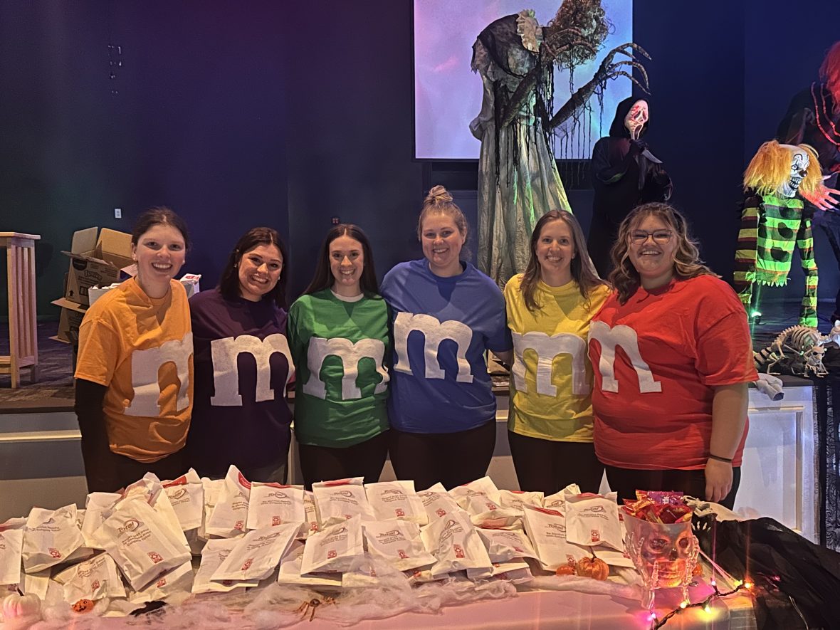 Dental club students wearing colorful M&M-inspired shirts stand behind a table filled with treat bags at a Halloween Trunk or Treat event.