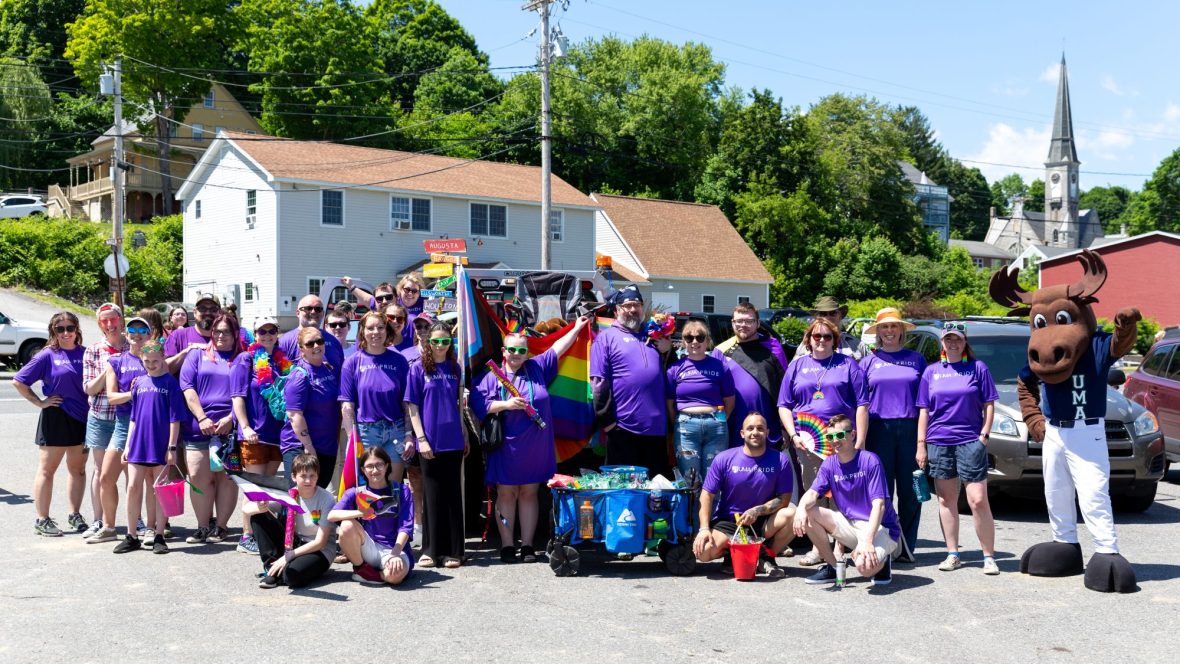 A group of people of diverse ages in colorful matching t-shirts pose with UMA Moose mascot Augustus at a pride parade in Hallowell, Maine