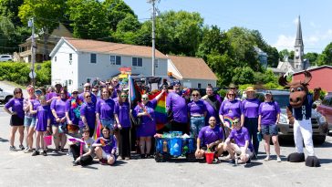 A group of people of diverse ages in colorful matching t-shirts pose with UMA Moose mascot Augustus at a pride parade in Hallowell, Maine