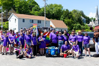 A group of people of diverse ages in colorful matching t-shirts pose with UMA Moose mascot Augustus at a pride parade in Hallowell, Maine