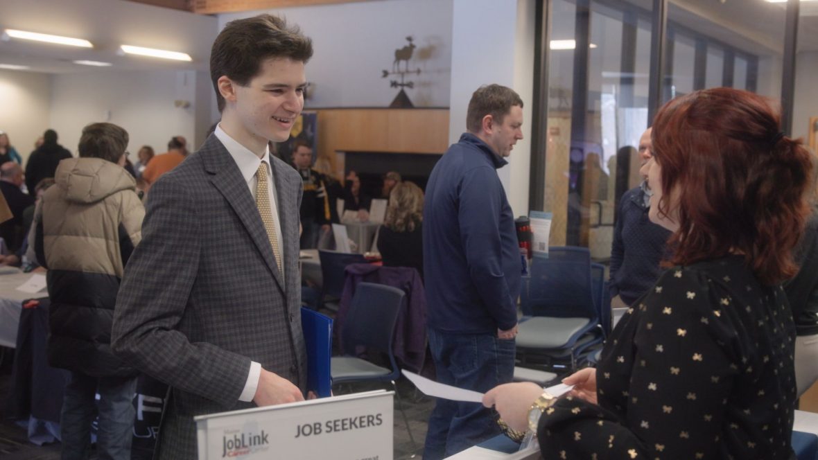 A student in a suit speaks with an employer across a table labeled “Job Seekers” during a busy job fair at the University of Maine at Augusta while attendees connect with recruiters and explore career opportunities.