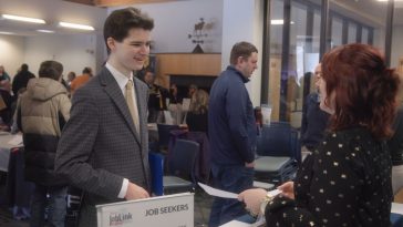 A student in a suit speaks with an employer across a table labeled “Job Seekers” during a busy job fair at the University of Maine at Augusta while attendees connect with recruiters and explore career opportunities.