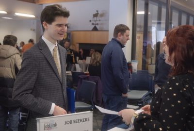A student in a suit speaks with an employer across a table labeled “Job Seekers” during a busy job fair at the University of Maine at Augusta while attendees connect with recruiters and explore career opportunities.