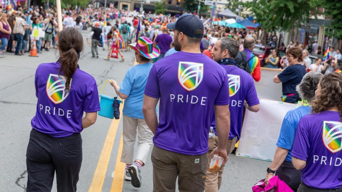 A group of people wearing purple shirts with the UMA rainbow shield logo and the word “PRIDE” walk together in a parade, facing a large crowd lining a city street filled with colorful outfits, flags, and tents.