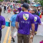 A group of people wearing purple shirts with the UMA rainbow shield logo and the word “PRIDE” walk together in a parade, facing a large crowd lining a city street filled with colorful outfits, flags, and tents.