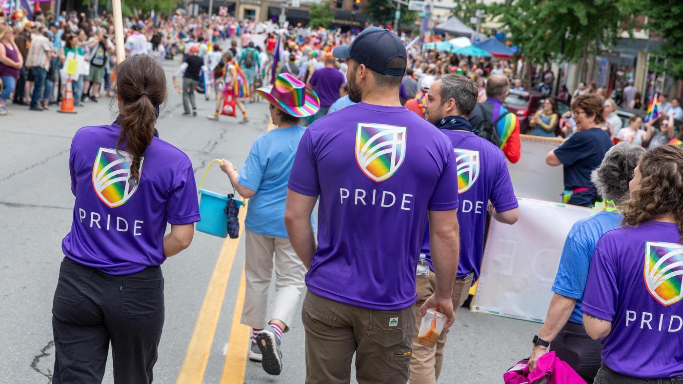 A group of people wearing purple shirts with the UMA rainbow shield logo and the word “PRIDE” walk together in a parade, facing a large crowd lining a city street filled with colorful outfits, flags, and tents.