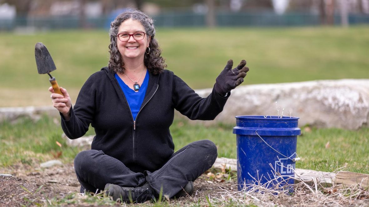 President Cushman sits cross-legged outdoors holding a hand trowel and wearing gardening gloves beside a blue bucket, with grass and a low stone wall in the background.