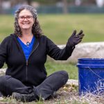 President Cushman sits cross-legged outdoors holding a hand trowel and wearing gardening gloves beside a blue bucket, with grass and a low stone wall in the background.