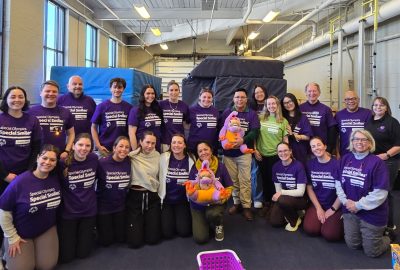 A group of dental hygiene students, faculty, and volunteers wearing matching purple Special Olympics shirts gather indoors for a group photo. Some kneel in the front row while others stand behind, smiling.