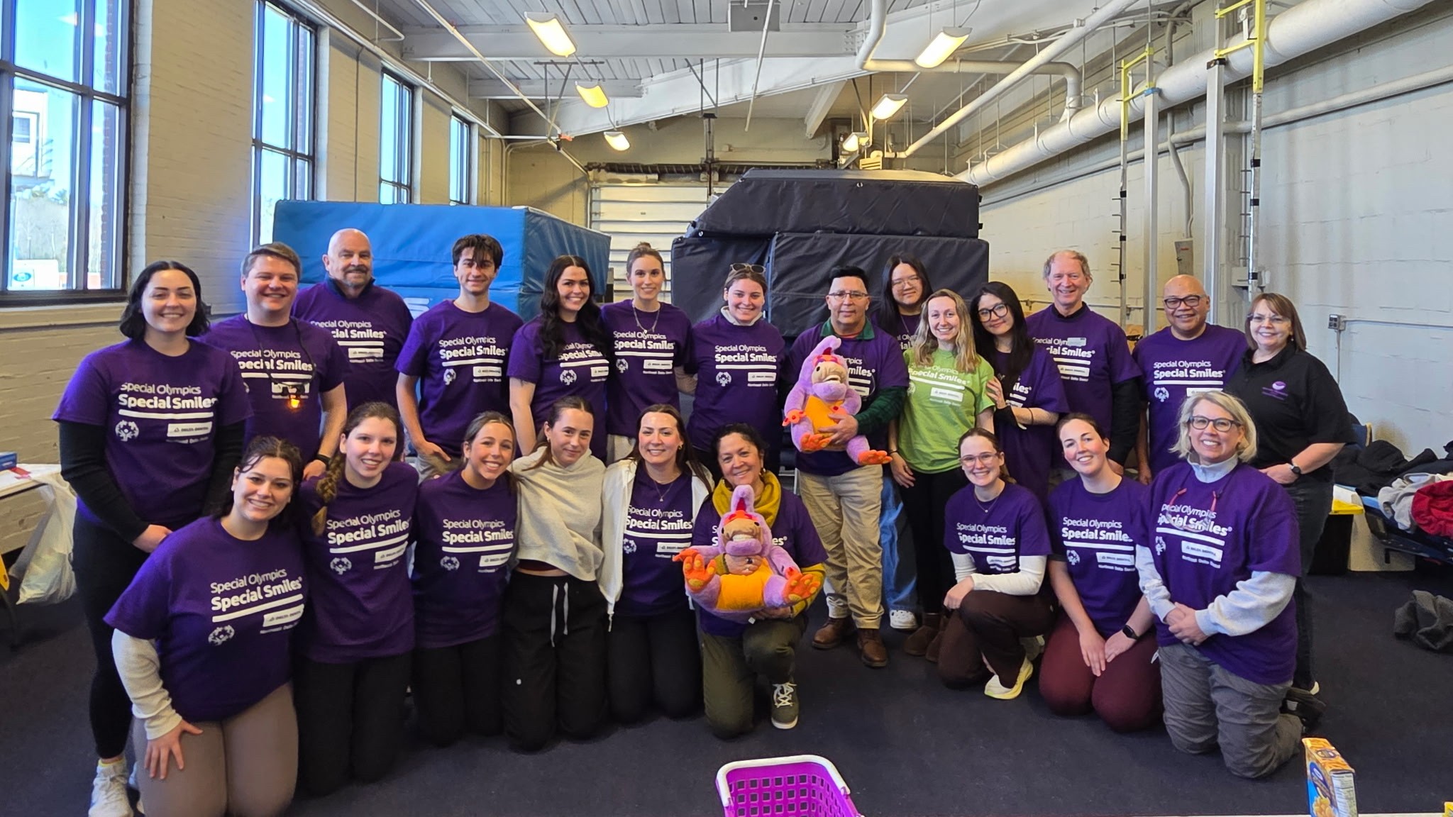 A group of dental hygiene students, faculty, and volunteers wearing matching purple Special Olympics shirts gather indoors for a group photo. Some kneel in the front row while others stand behind, smiling. 