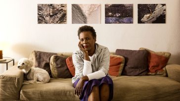 Author Claudia Rankine sits on a couch, elbow on knee and chin resting on hand, her miniature poodle rests its head on the arm of the couch.