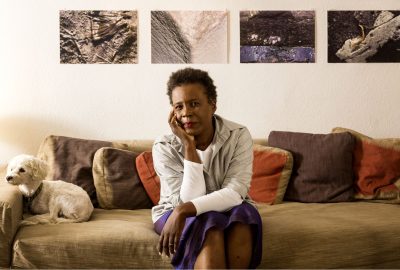 Author Claudia Rankine sits on a couch, elbow on knee and chin resting on hand, her miniature poodle rests its head on the arm of the couch.