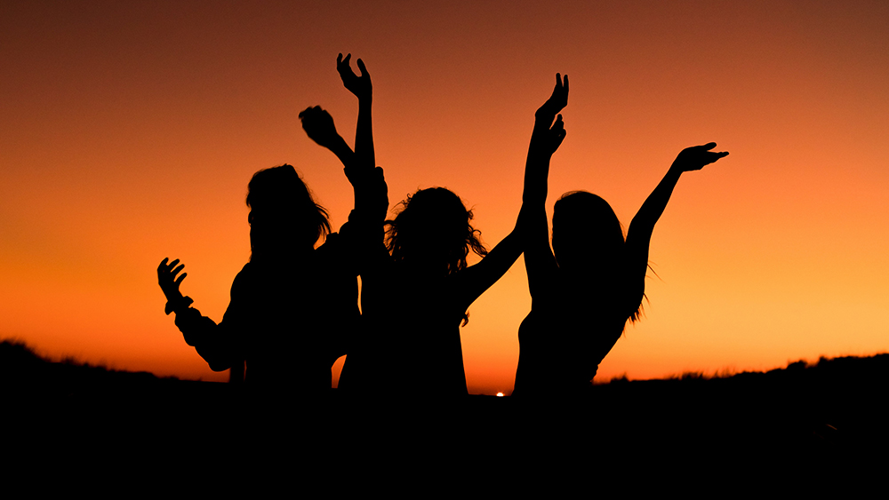 Silhouettes of three people standing with their arms raised against a bright orange sunset sky.