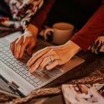 Person typing on a laptop while sitting on a bed or couch, wrapped in a patterned blanket, with a mug and notebook nearby in a cozy setting.