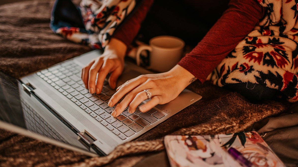 Person typing on a laptop while sitting on a bed or couch, wrapped in a patterned blanket, with a mug and notebook nearby in a cozy setting.