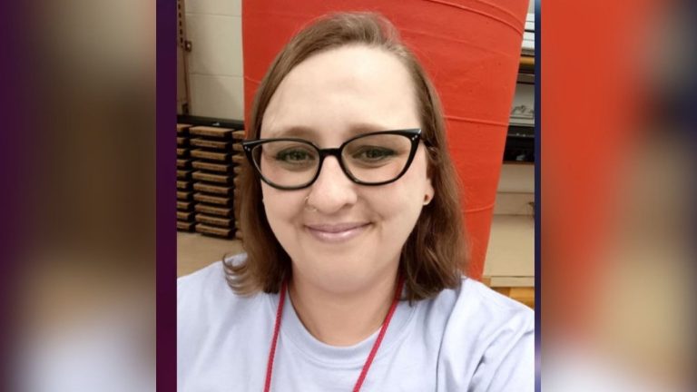 Rachel Dollahan, a smiling woman wearing glasses, a delicate nose ring and small red earrings, dressed in a tshirt in front of a red background.