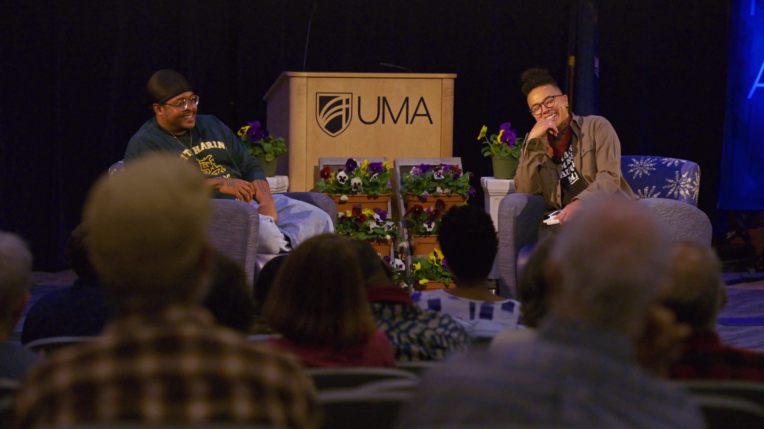 Two poets, Danez Smith and Ian-Khara Ellasante, sit on stage in armchairs, smiling and laughing during a conversation. A wooden podium with the UMA logo stands behind and between them, decorated with small arrangements of purple and yellow flowers. The audience is visible in the foreground, slightly out of focus, as they watch the exchange.