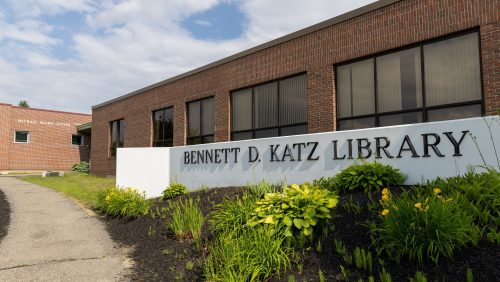 View of the Bennett D. Katz Library building at the University of Maine at Augusta, with its large sign and landscaped entrance in the foreground and the attached red-brick Michael Klahr Center visible to the left in the background.