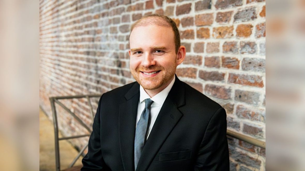 Chandler Dugal, wearing a suit and tie smiles while seated on a metal bench in front of a brick wall