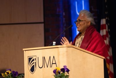Claudia Rankine stands at a wooden podium bearing the UMA logo, smiling as she speaks into a microphone. She wears glasses and a red shawl draped over her shoulders. A small bouquet of purple and yellow flowers sits at the front of the podium. The background includes a softly lit stage, a blue accent light, and a partially visible American flag.