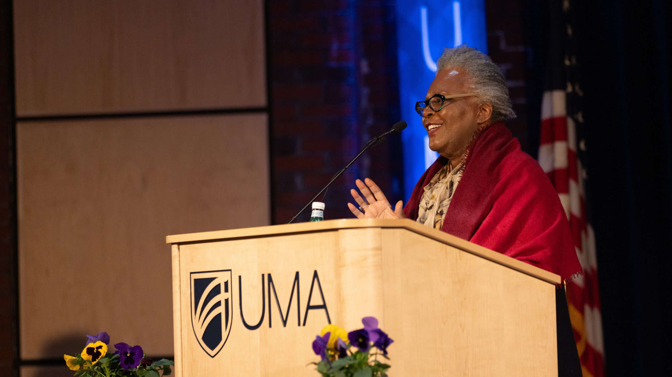 Claudia Rankine stands at a wooden podium bearing the UMA logo, smiling as she speaks into a microphone. She wears glasses and a red shawl draped over her shoulders. A small bouquet of purple and yellow flowers sits at the front of the podium. The background includes a softly lit stage, a blue accent light, and a partially visible American flag.