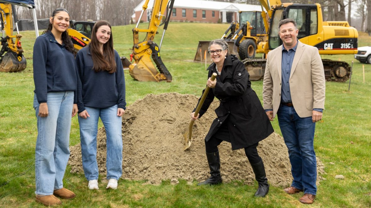 Four people stand on a grassy site during a groundbreaking ceremony, with UMA President Jenifer Cushman shoveling dirt from a small mound while Lilian Cox, Skyla Dean and Matt Morrill stand beside her. Excavators and a brick building are visible in the background.
