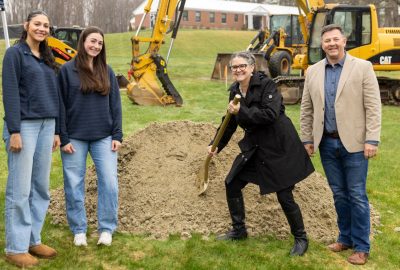 Four people stand on a grassy site during a groundbreaking ceremony, with UMA President Jenifer Cushman shoveling dirt from a small mound while Lilian Cox, Skyla Dean and Matt Morrill stand beside her. Excavators and a brick building are visible in the background.