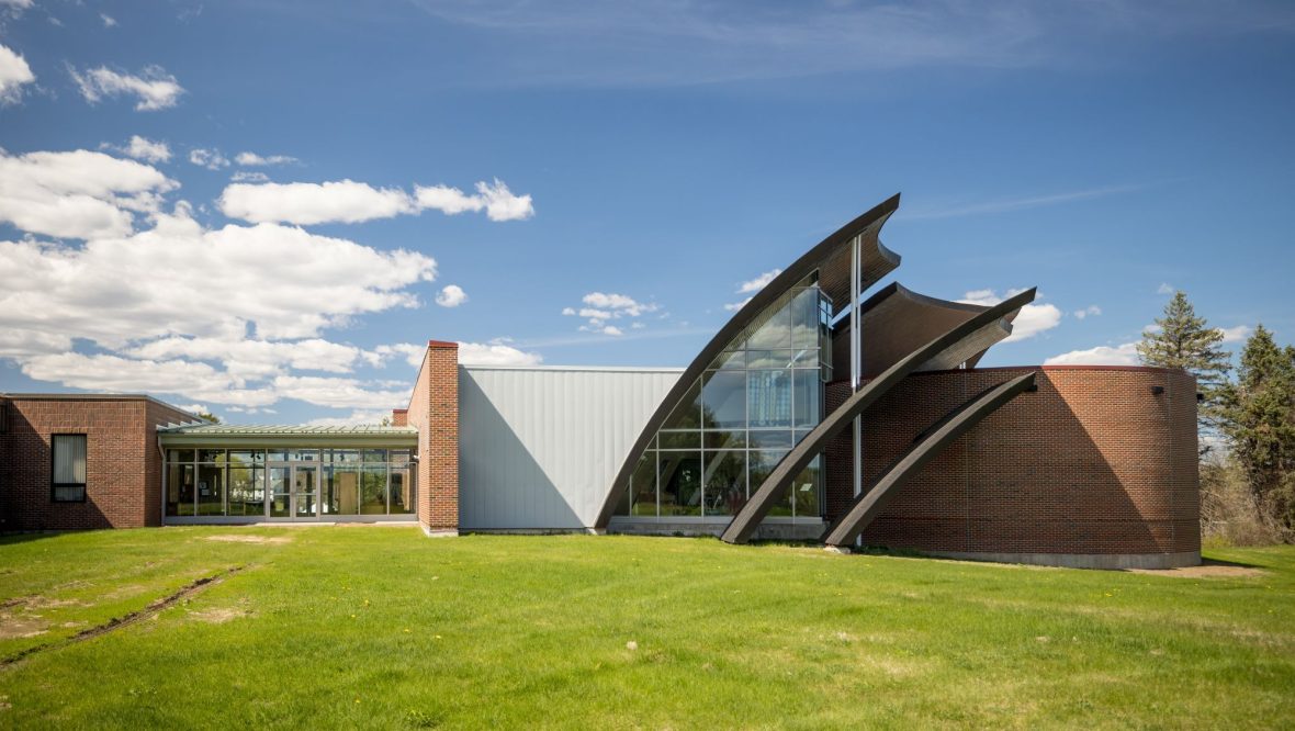 Wide view of the Michael Klahr Center at the University of Maine at Augusta, showing its curved, petal-like glass and brick façade set against a blue sky and open lawn.