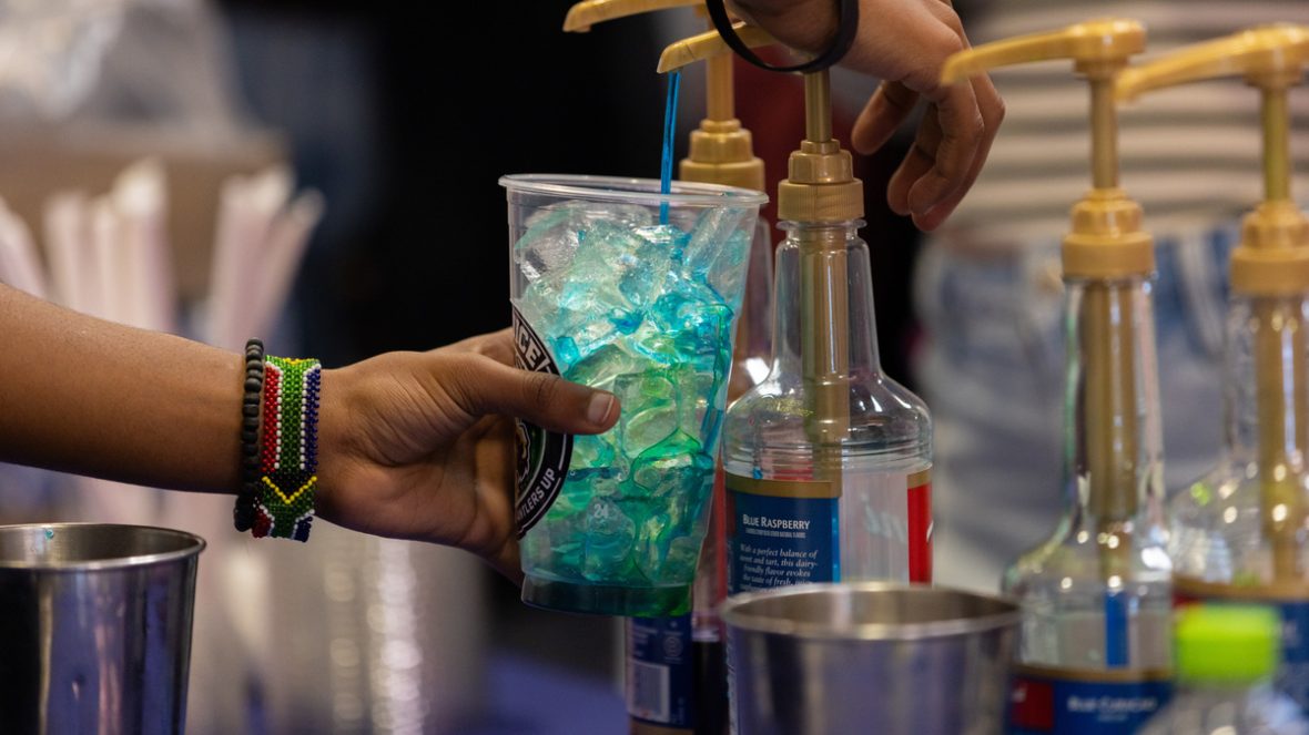 A person fills a clear plastic cup with bright blue syrup from a pump bottle, pouring it over ice while another hand holds the cup steady; several flavored syrup bottles and metal containers sit nearby on a counter.