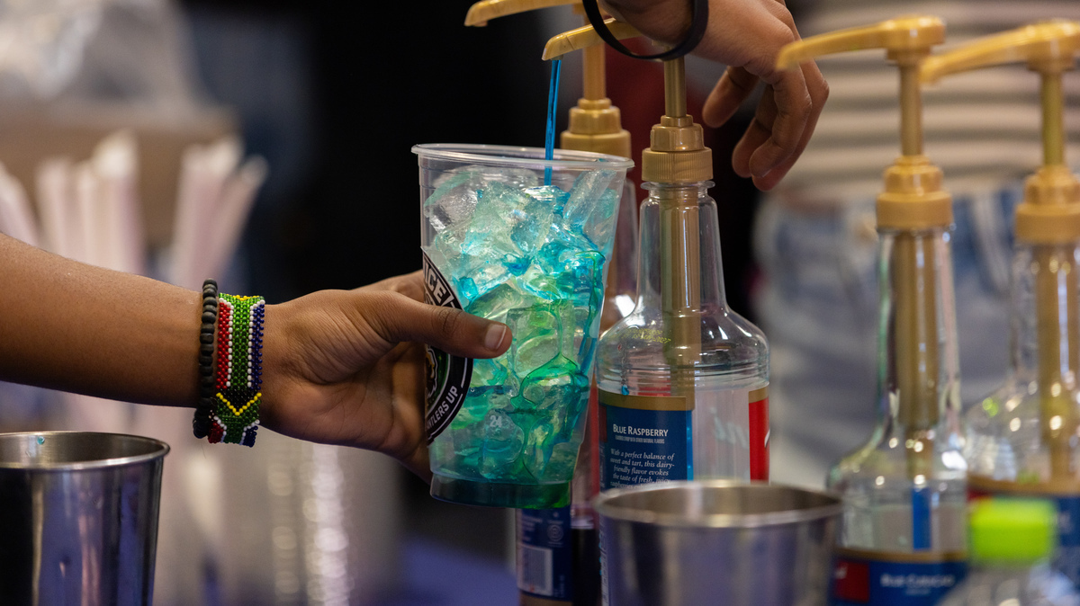 A person fills a clear plastic cup with bright blue syrup from a pump bottle, pouring it over ice while another hand holds the cup steady; several flavored syrup bottles and metal containers sit nearby on a counter.