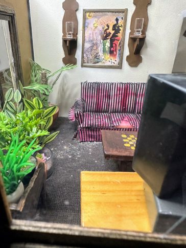 Still life of a dollhouse living room with plants, a worn striped couch, wall sconces, painting, table and the back of a television in the foreground.