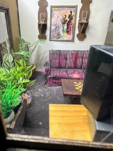 Still life of a dollhouse living room with plants, a worn striped couch, wall sconces, painting, table and the back of a television in the foreground.