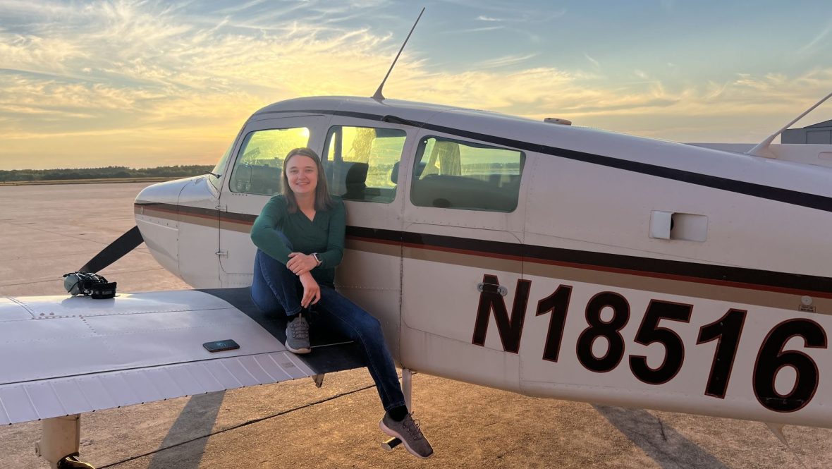 A woman sits on the wing of a small airplane at an airport during sunset, smiling beside the aircraft under a colorful sky.