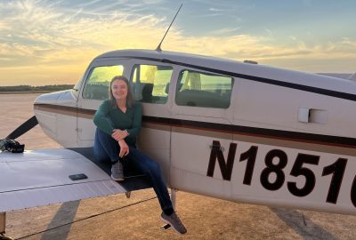 A woman sits on the wing of a small airplane at an airport during sunset, smiling beside the aircraft under a colorful sky.