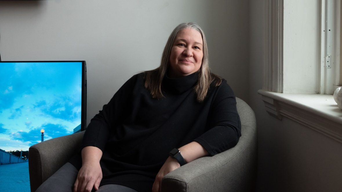Bethany Engstrom is seated on a u-shaped chair next to a window to her left. A large piece of art sits on the floor to her right leaning against the wall behind her.