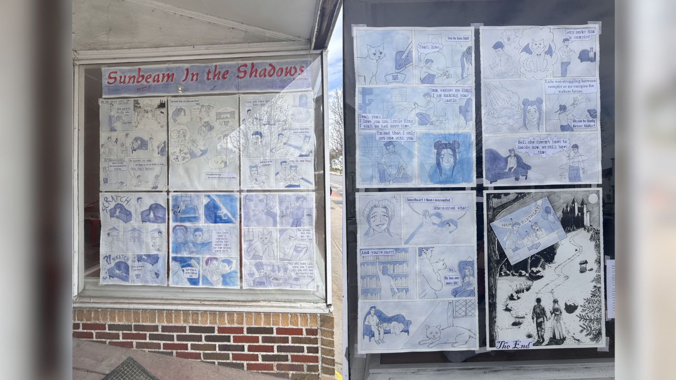Storefront gallery windows displaying student comic and sequential art projects from a Graphic Storytelling course at Art:Works on Main in Winthrop. The installation includes hand-drawn narrative panels and illustrated scenes arranged across two large window displays.