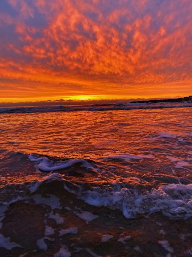 A vivid orange and purple sunset over the ocean with waves lapping at the shore.